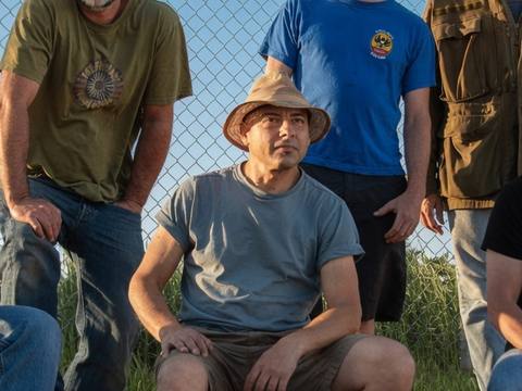 Six band members sitting against a chain-link fence under blue sky, casual promo shot